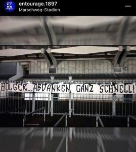 Auf dem Bild zu sehen: Ein Spruchband hängt an Gittern vor einem Stadion. Darauf die Aufschrift: " Holger ... Abdanken, ganz Schnell!".